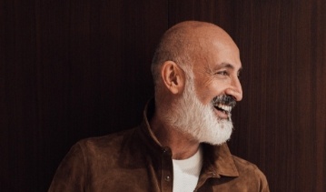 Older man with white beard standing in front of dark wood wall