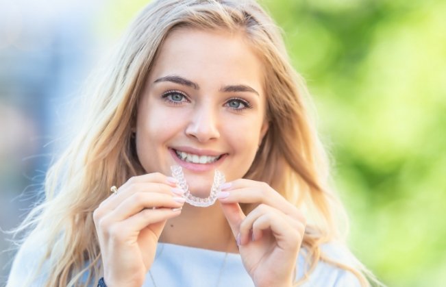 Smiling blonde woman holding SureSmile clear aligner outdoors