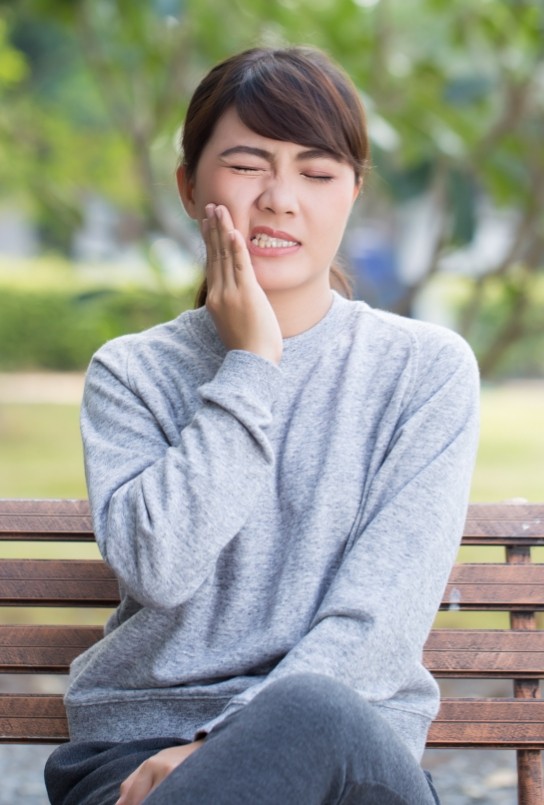 Woman on park bench holding cheek in pain needing wisdom tooth extractions in Covington